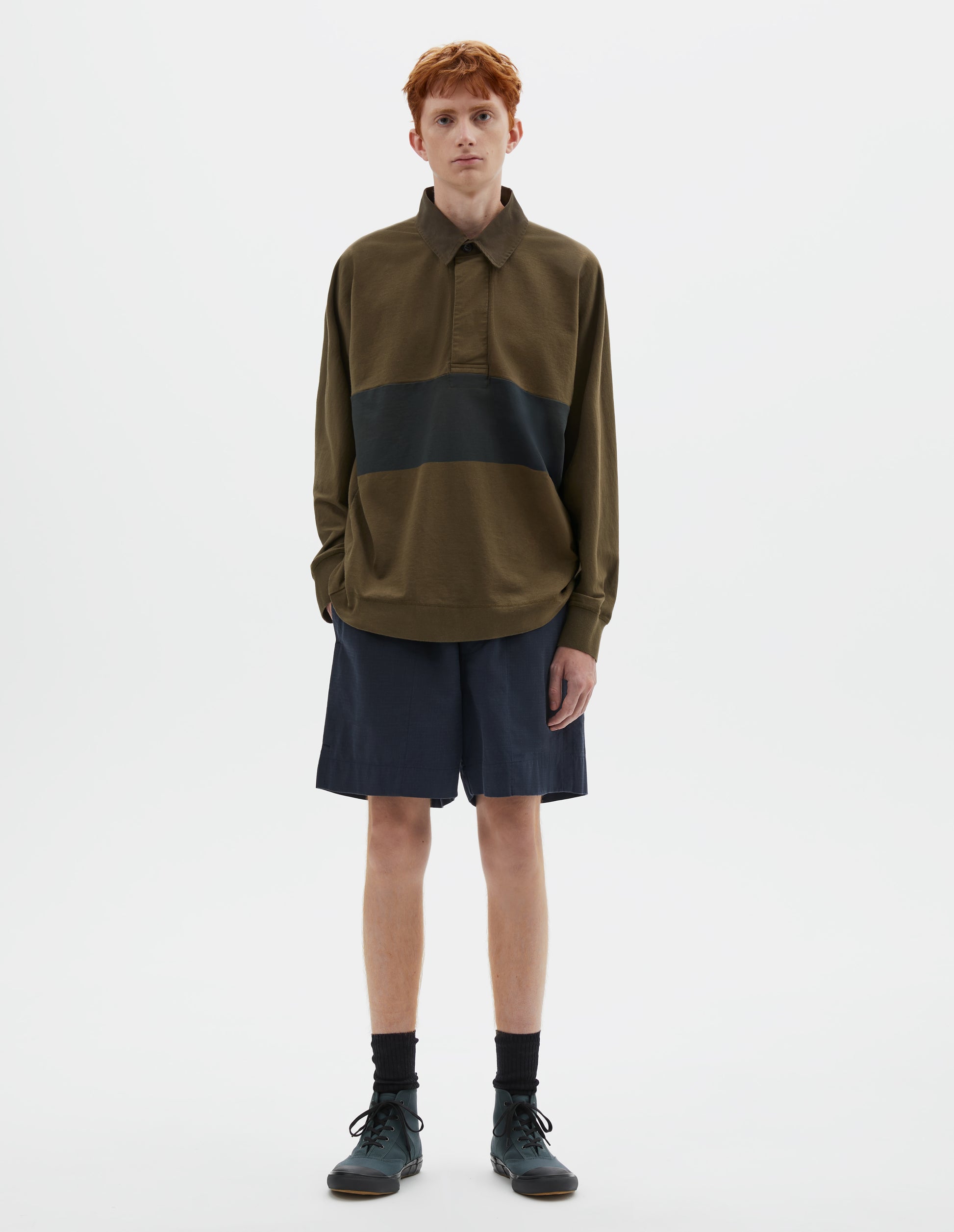 Full body image, front view of model wearing dark green rugby long sleeve polo shirt with black panel across chest, twill collar, wide fly front placket and rib cuffs. Styled with navy blue shorts, black socks and blue canvas trainers.