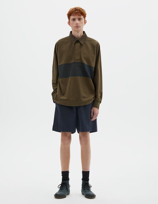 Full body image, front view of model wearing dark green rugby long sleeve polo shirt with black panel across chest, twill collar, wide fly front placket and rib cuffs. Styled with navy blue shorts, black socks and blue canvas trainers.