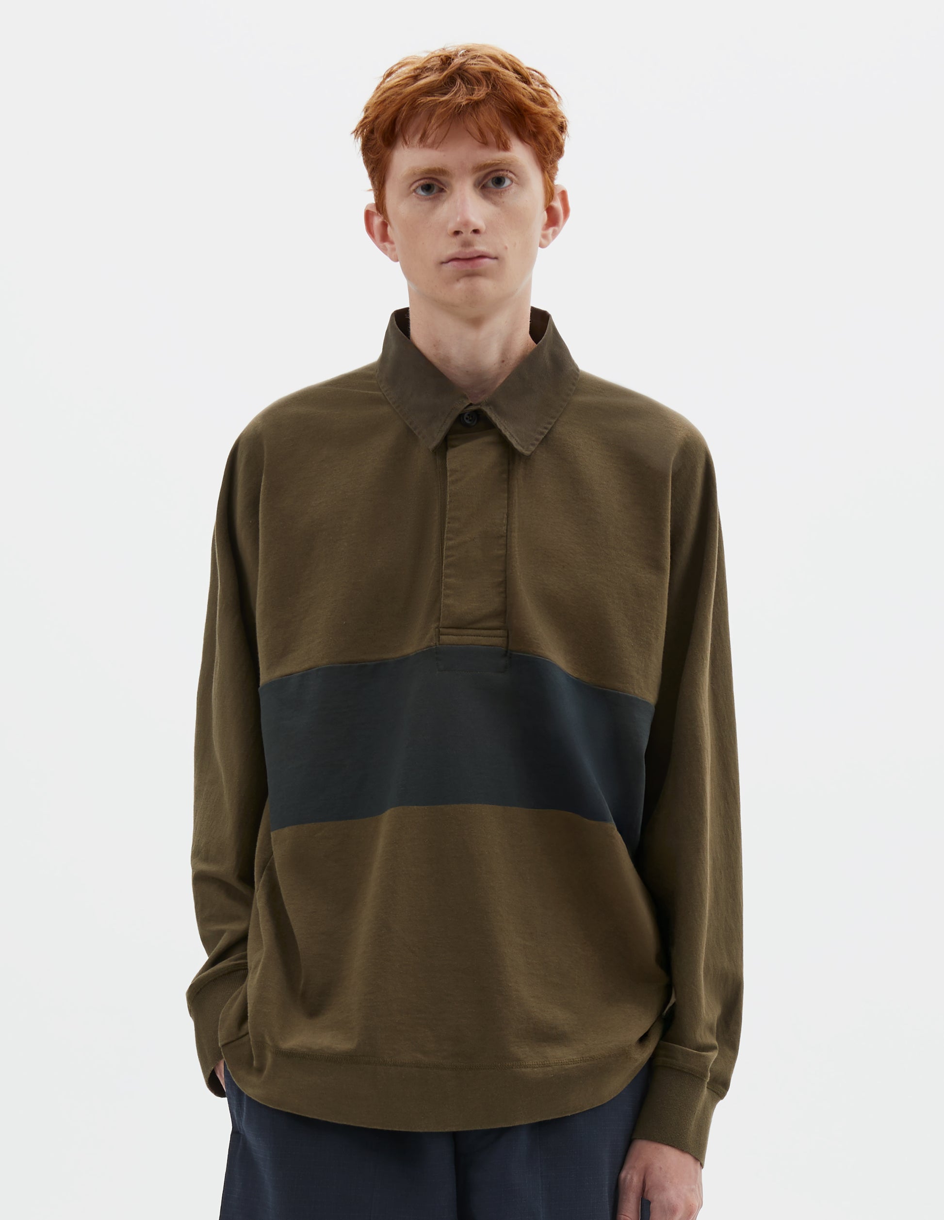 Cropped body image, front view of model wearing dark green rugby long sleeve polo shirt with black panel across chest, twill collar, wide fly front placket and rib cuffs. Styled with navy blue shorts.