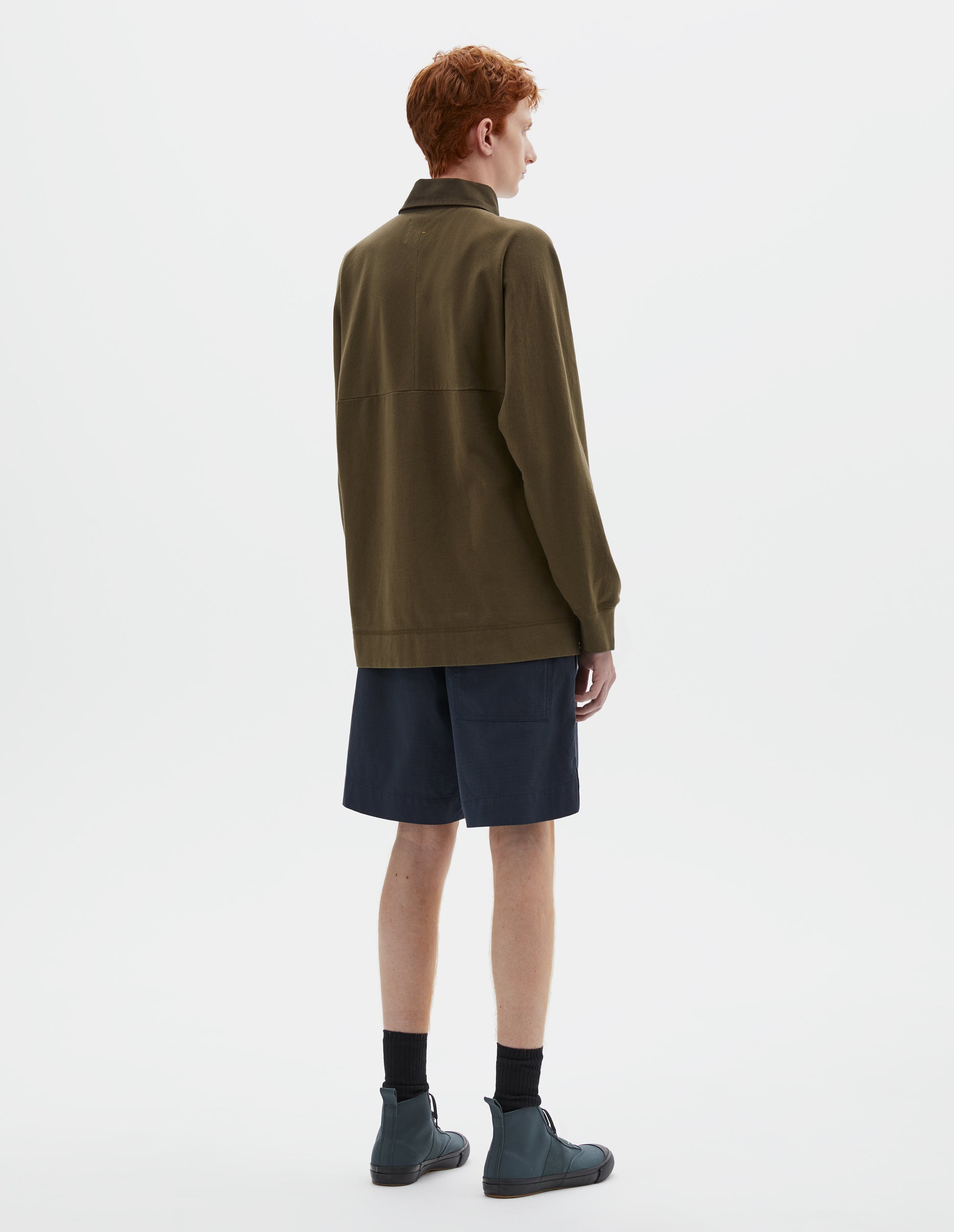 Full body image, back view of model wearing dark green rugby long sleeve polo shirt with twill collar and rib cuffs. Styled with navy blue shorts, black socks and blue canvas trainers.