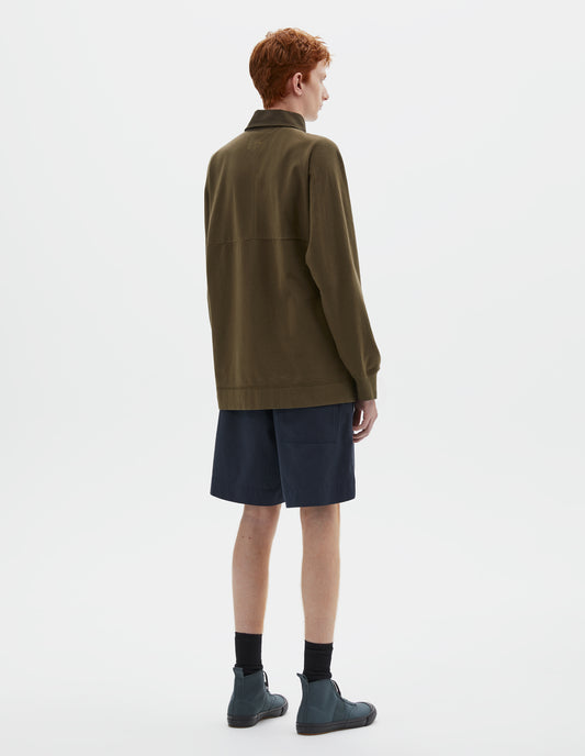 Full body image, back view of model wearing dark green rugby long sleeve polo shirt with twill collar and rib cuffs. Styled with navy blue shorts, black socks and blue canvas trainers.