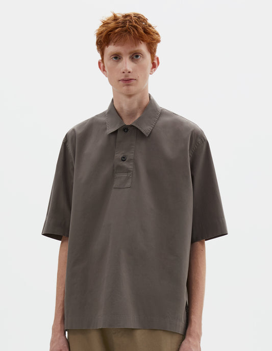 Cropped body image, front view of model wearing relaxed greyish brown short-sleeved shirt with corozo brace buttons on wide offset placket and vent split hem. Styled with dark beige trousers.