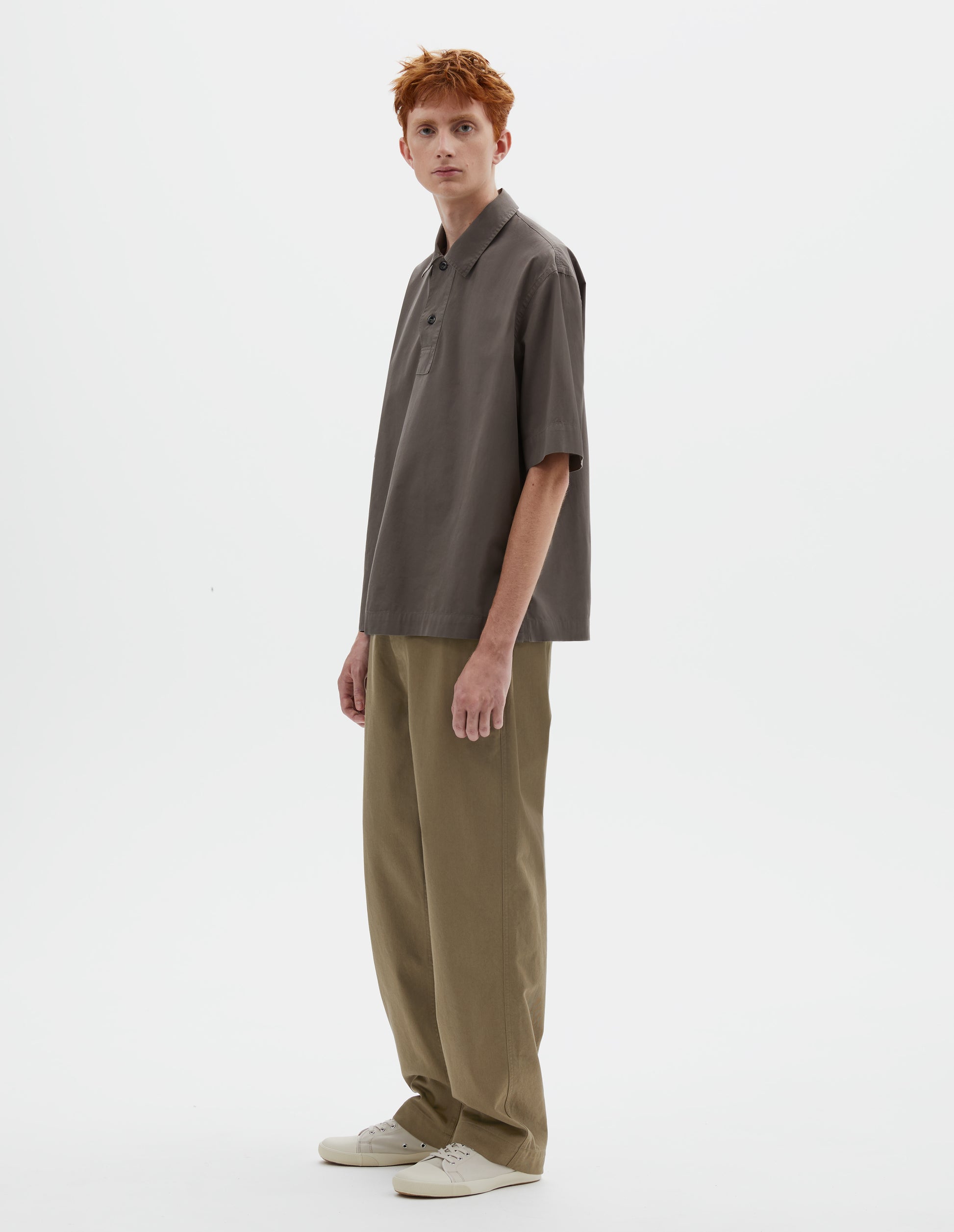 Full body image, front view of model wearing relaxed greyish brown short-sleeved shirt with corozo brace buttons on wide offset placket and vent split hem. Styled with dark beige trousers and light beige canvas trainers.
