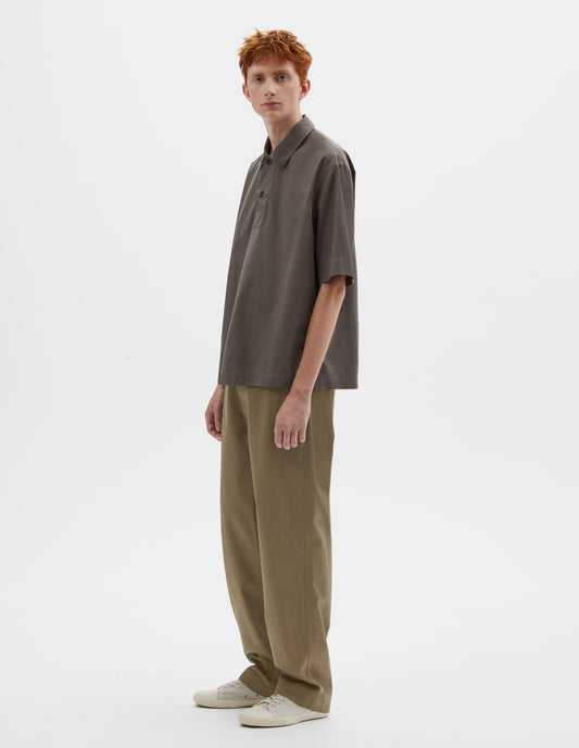 Full body image, front view of model wearing relaxed greyish brown short-sleeved shirt with corozo brace buttons on wide offset placket and vent split hem. Styled with dark beige trousers and light beige canvas trainers.
