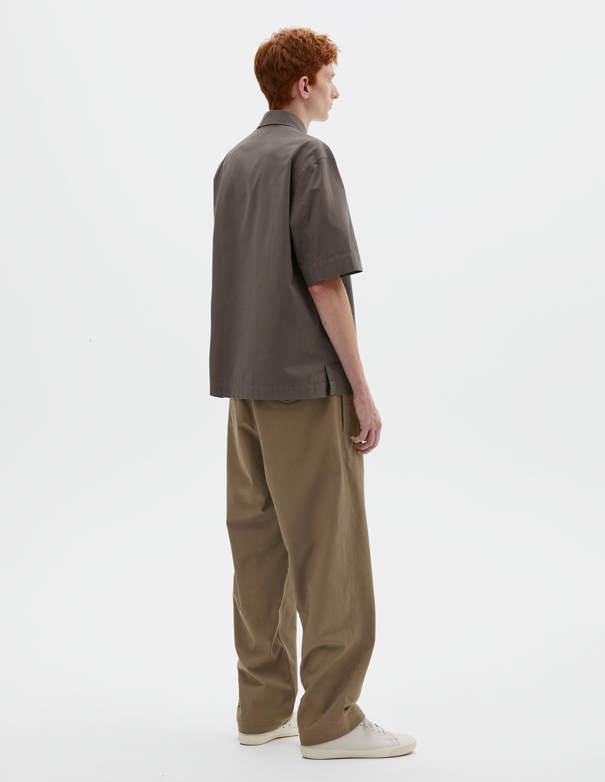 Full body image, back view on  model green cotton drill trousers with belt loops, side adjustors, welt back and flap back pockets. Styled with grey short sleeve shirt and beige canvas trainers.