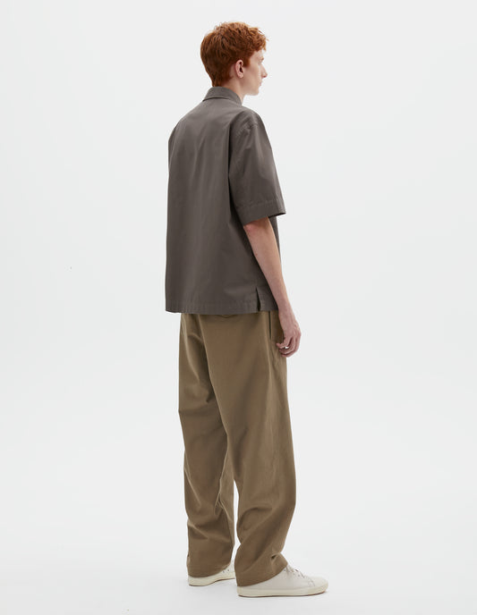 Full body image, back view on  model green cotton drill trousers with belt loops, side adjustors, welt back and flap back pockets. Styled with grey short sleeve shirt and beige canvas trainers.