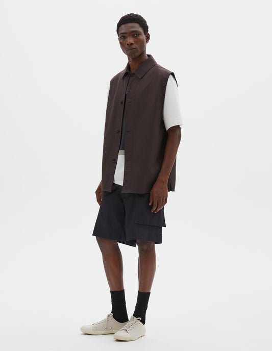 Full body image, front view of model wearing dark brown cotton drill sleeveless overshirt with corozo button fastening and vent split hem. Styled over white t-shirt, black cargo shorts and black socks with beige plimsolls.