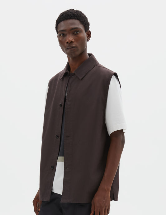 Cropped body image, front view of model wearing dark brown cotton drill sleeveless overshirt with corozo button fastening and vent split hem. Styled over white t-shirt and black cargo shorts.