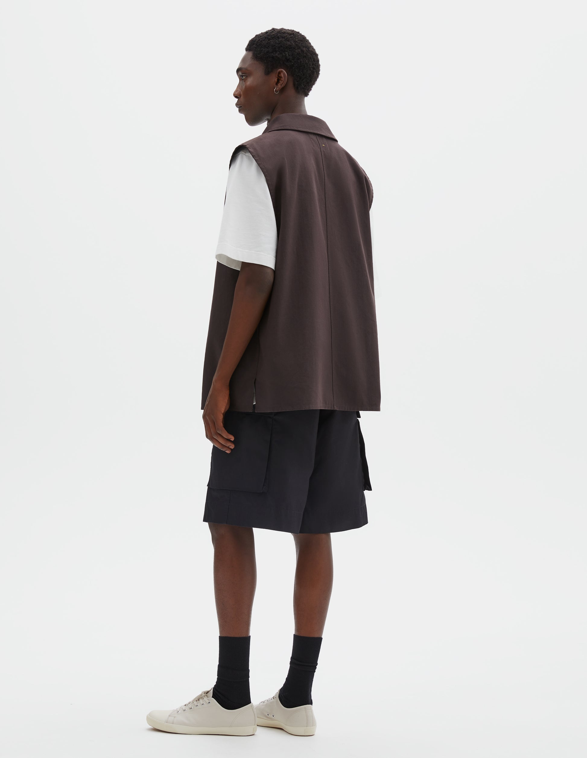 Full body image, back view of model wearing dark brown cotton drill sleeveless overshirt, with centre seam detailing down back and vent split hem. Styled over white t-shirt, black cargo shorts and black socks with beige plimsolls.