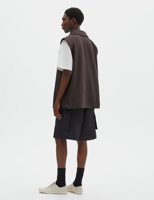 Full body image, back view of model wearing dark brown cotton drill sleeveless overshirt, with centre seam detailing down back and vent split hem. Styled over white t-shirt, black cargo shorts and black socks with beige plimsolls.