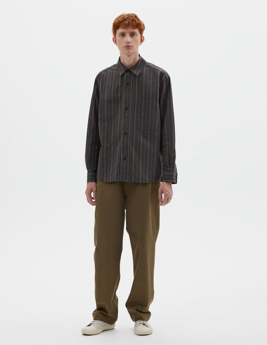 Full body image, front view of model wearing off black and khaki striped shirt, with corozo button fastening and patch chest pocket. Styled with khaki green trousers and beige canvas trainers.