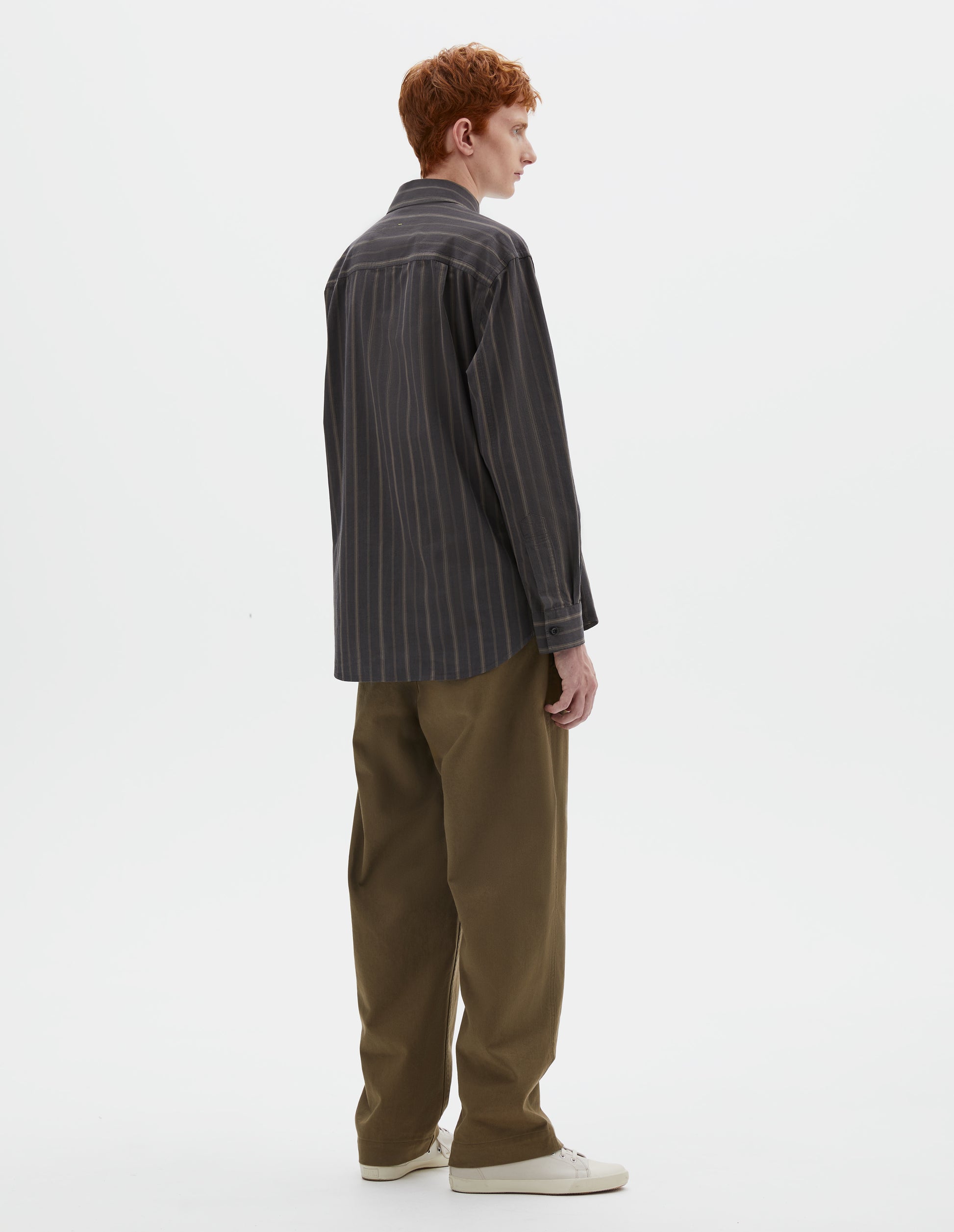 Full body image, back view of model wearing off black and khaki striped shirt, with corozo button cuffs and elongated curved hem. Styled with khaki green trousers and beige canvas trainers.