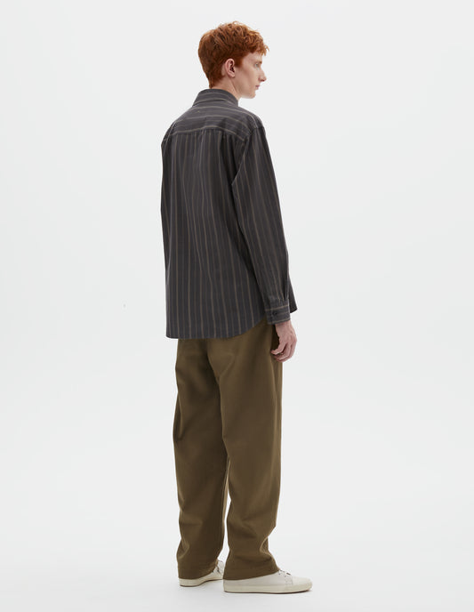 Full body image, back view of model wearing off black and khaki striped shirt, with corozo button cuffs and elongated curved hem. Styled with khaki green trousers and beige canvas trainers.