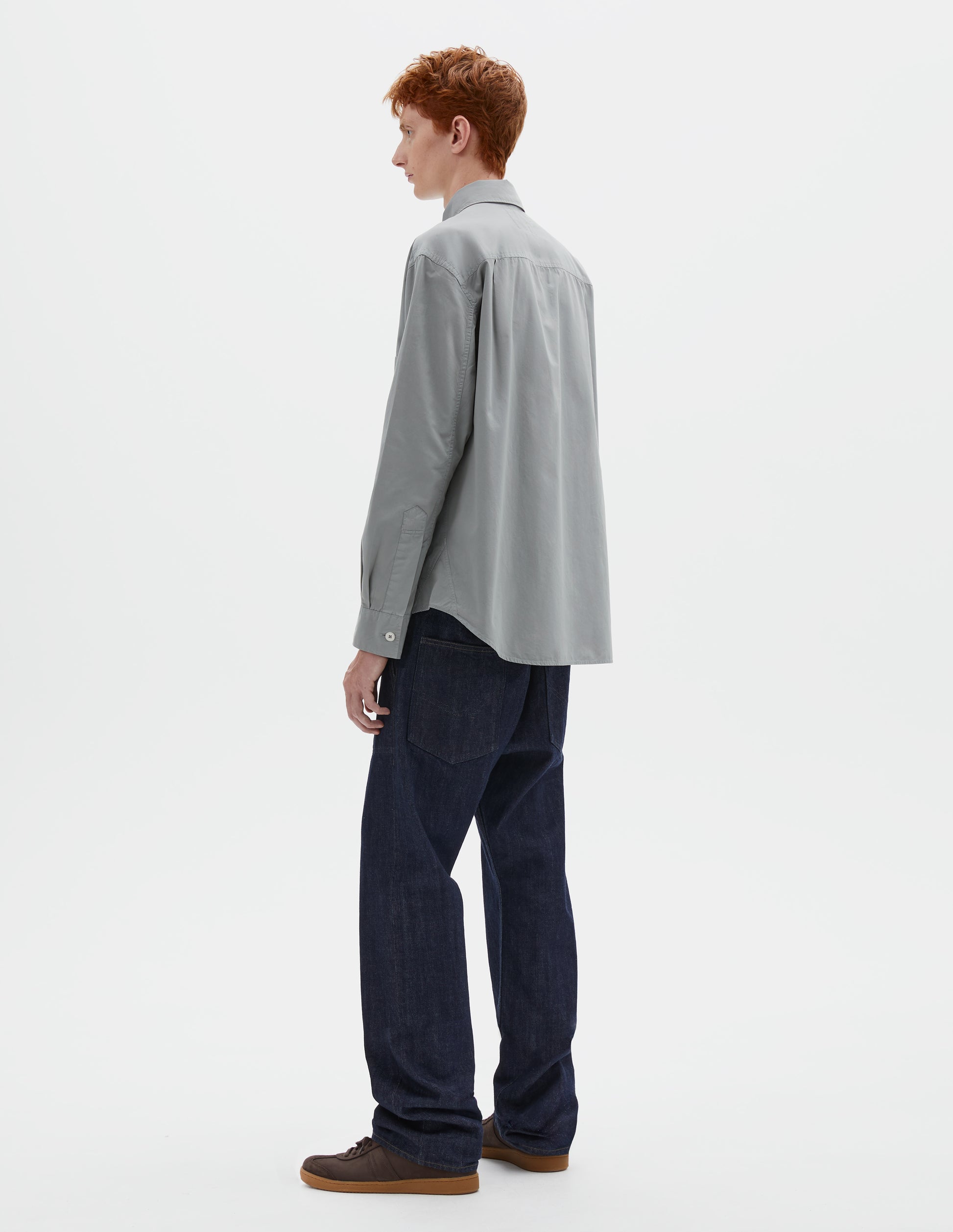 Full body image, front view of model wearing indigo denim twill jeans, with metal button fly, grown on waistband with belt loops and back pockets with half cotton lining. Styled with grey shirt and brown suede trainers.