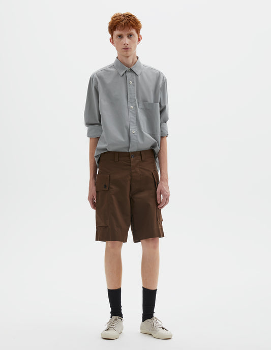Full body image, front view of model wearing brown cotton shorts with cargo pockets, belt loops and corozo brace button closure. Styled with greyish blue shirt tucked in, black socks and off white canvas trainers.