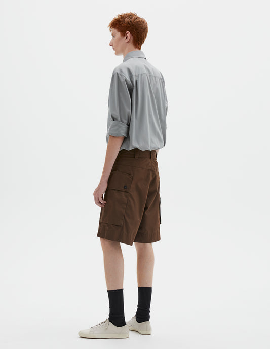Full body image, back view of model wearing brown cotton shorts with cargo pockets, belt loops and welt back pocket. Styled with greyish blue shirt tucked in, black socks and off white canvas trainers.