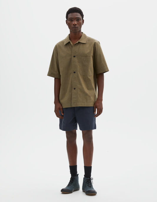 Full body image, front view of model wearing green cotton short sleeve shirt, with large patch chest pocket, side split hem and corozo brace button closure. Styled with blue shorts, black ankle length socks and blue canvas high top trainers.