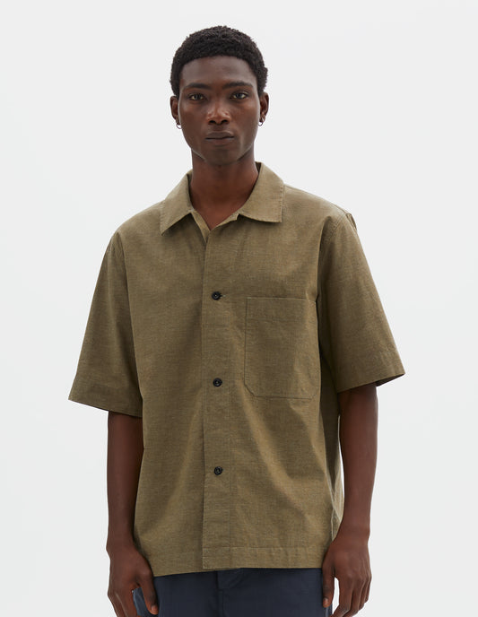 Cropped body image, front view of model wearing green cotton short sleeve shirt, with large patch chest pocket, side split hem and corozo brace button closure. Styled with blue shorts.