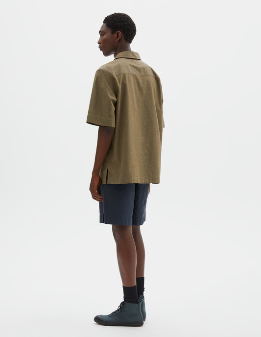 Full body image, back view of model wearing green cotton short sleeve shirt, with side split hem with contrasting herringbone tape. Styled with blue shorts, black ankle length socks and blue canvas high top trainers.