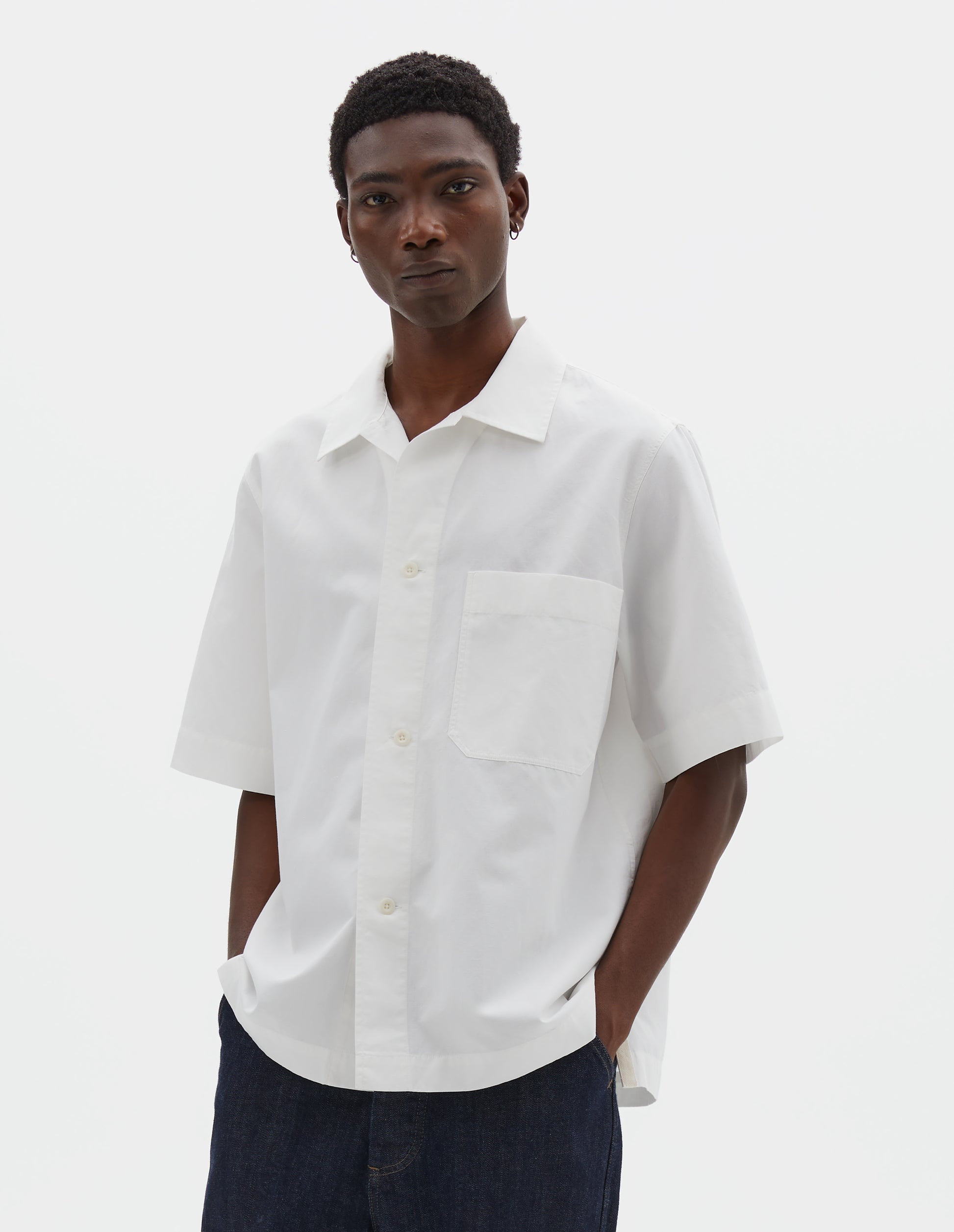 Cropped body image, front view of model wearing white cotton and linen short sleeve shirt, with large patch chest pocket, side split hem and corozo brace button closure. Styled with indigo blue jeans.