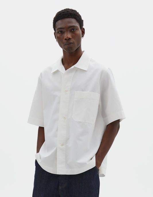 Cropped body image, front view of model wearing white cotton and linen short sleeve shirt, with large patch chest pocket, side split hem and corozo brace button closure. Styled with indigo blue jeans.