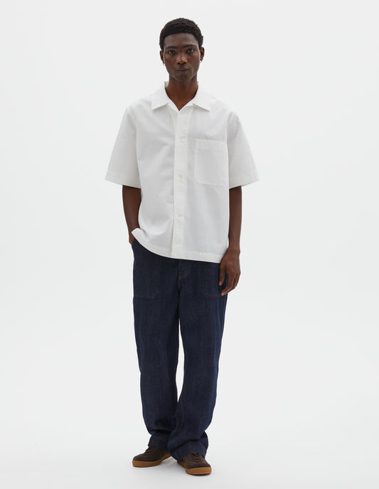 Full body image, front view of model wearing white cotton and linen short sleeve shirt, with large patch chest pocket, side split hem and corozo brace button closure. Styled with indigo blue jeans and brown suede trainers.