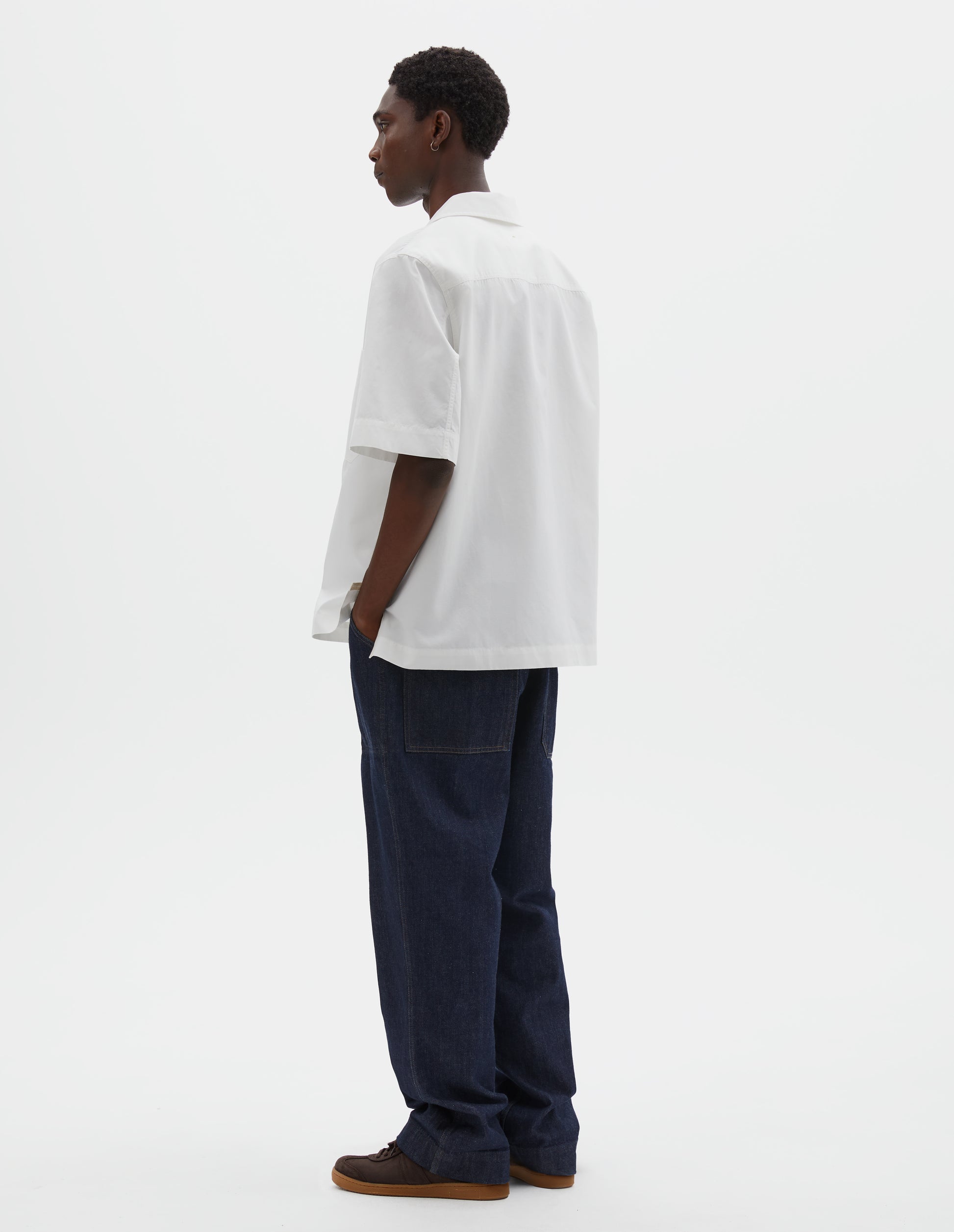 Full body image, back view of model wearing indigo blue denim twill jeans. With metal button tab waist adjusters, belt loops and patch and hammer pocket on back of trousers. Styled with white shirt and brown suede trainers.