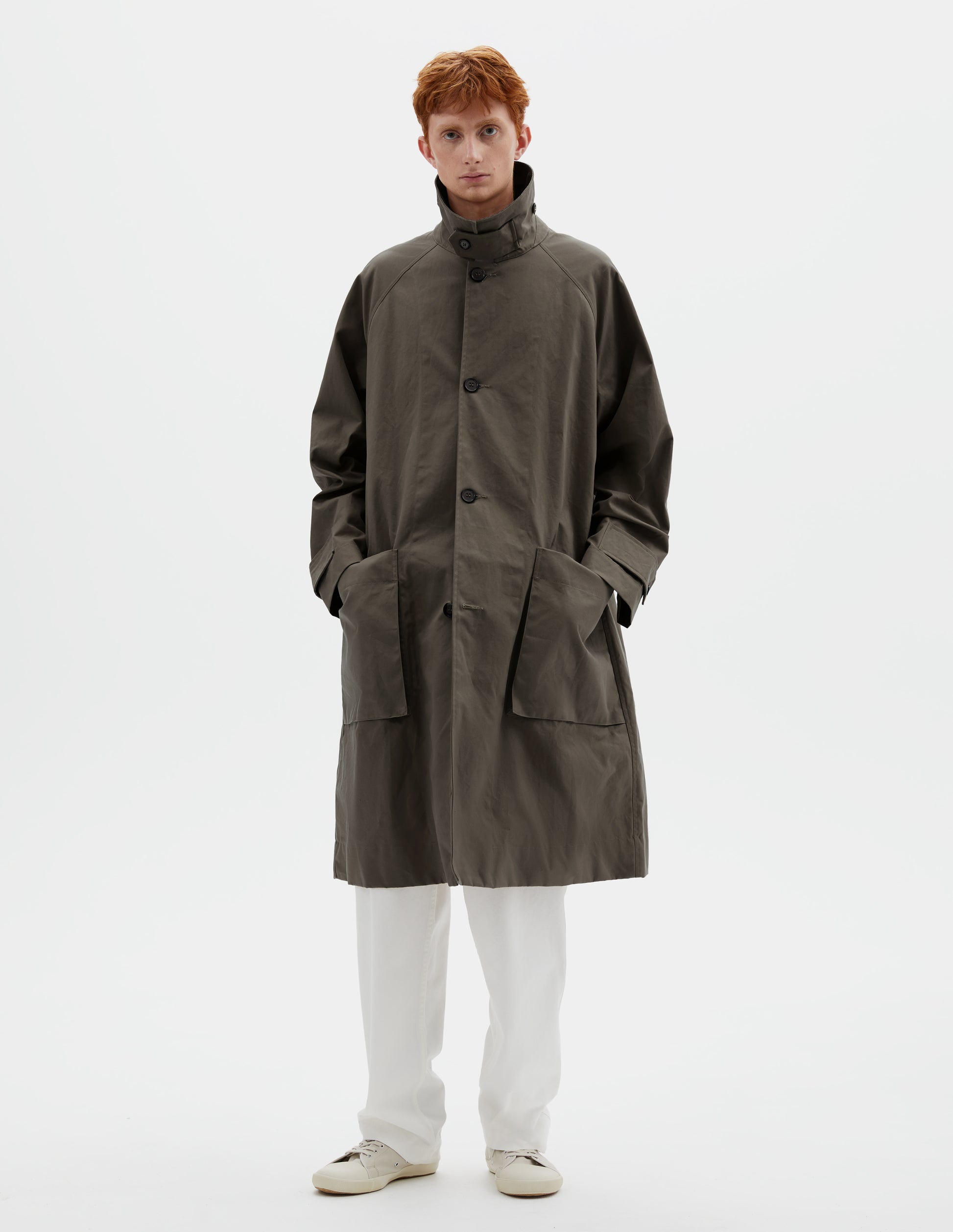 Full body image, front view of model wearing greyish brown long sleeve cotton mac, with storm tab fastening neck, raglan sleeve, button tab adjustable cuffs and large patch bellow pockets. Styled with collar popped, over white cotton trousers and beige canvas trainers.