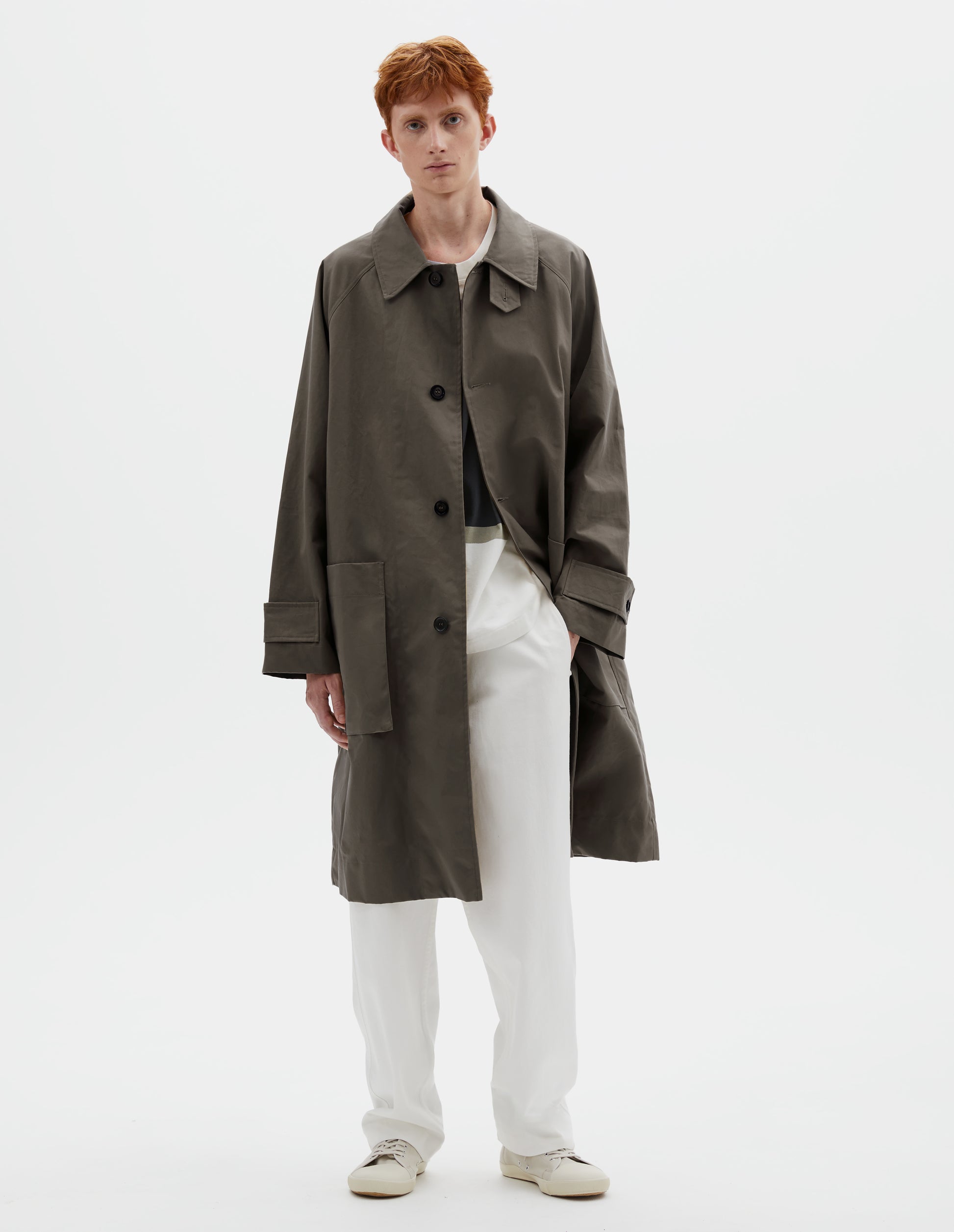 Full body image, front view of model wearing greyish brown long sleeve cotton mac, with storm tab fastening neck, raglan sleeve, button tab adjustable cuffs and large patch bellow pockets. Styled open, over white cotton t-shirt with white cotton trousers and beige canvas trainers.