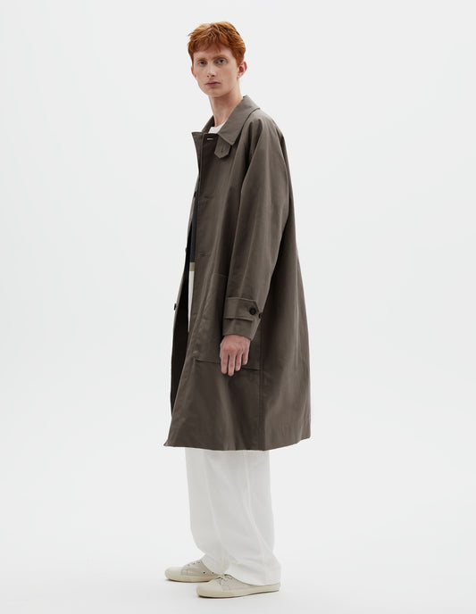 Full body image, side view of model wearing greyish brown long sleeve cotton mac, with storm tab fastening neck, raglan sleeve, button tab adjustable cuffs and large patch bellow pockets. Styled with white cotton trousers and beige canvas trainers.