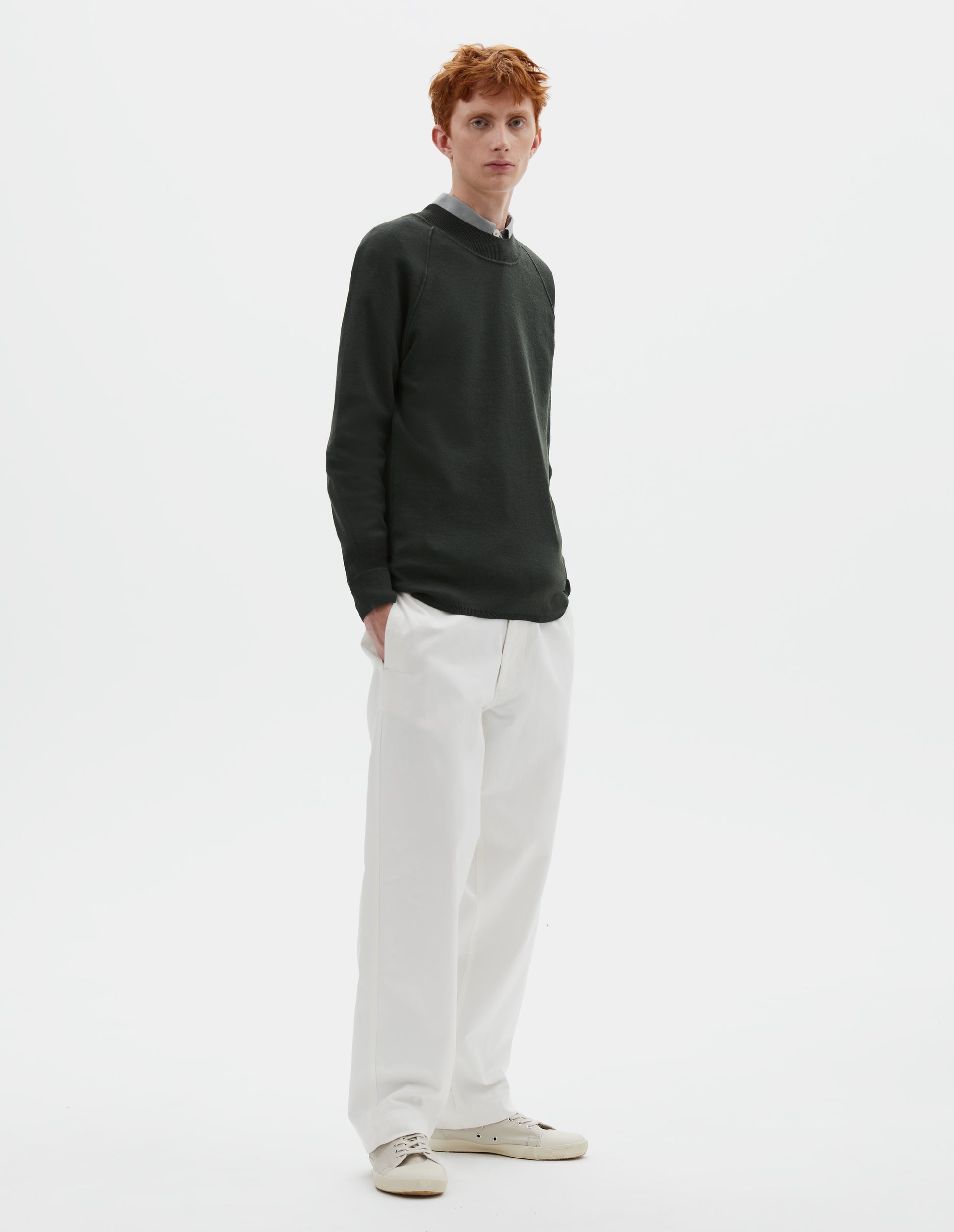Full body image, front view of model wearing dark grey merino wool sweatshirt with raglan sleeve and contrast rib on neck, cuffs and hem. Styled over grey shirt, with white trousers and beige canvas trainers.