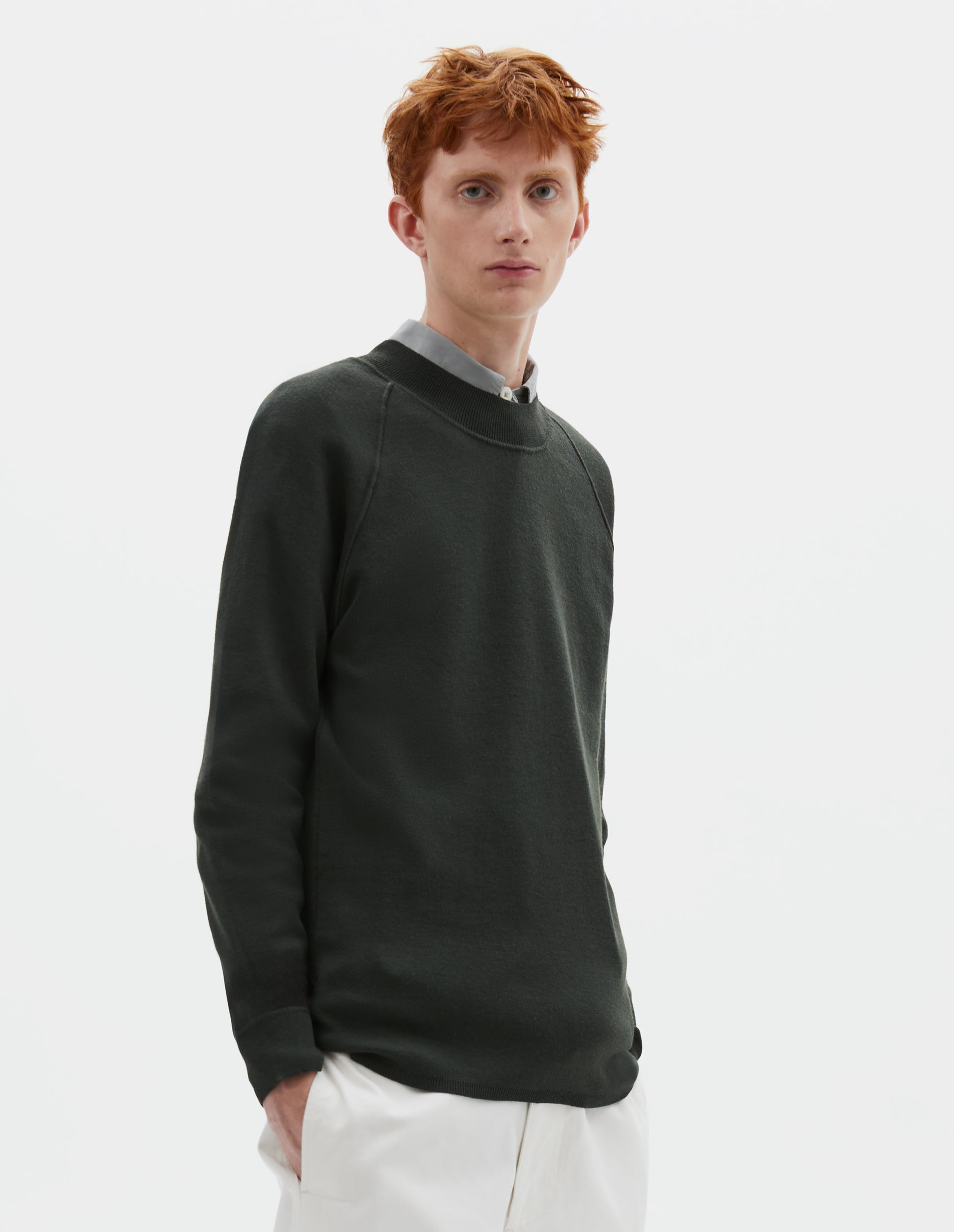 Cropped body image, front view of model wearing dark grey merino wool sweatshirt with raglan sleeve and contrast rib on neck, cuffs and hem. Styled over grey shirt, with white trousers.