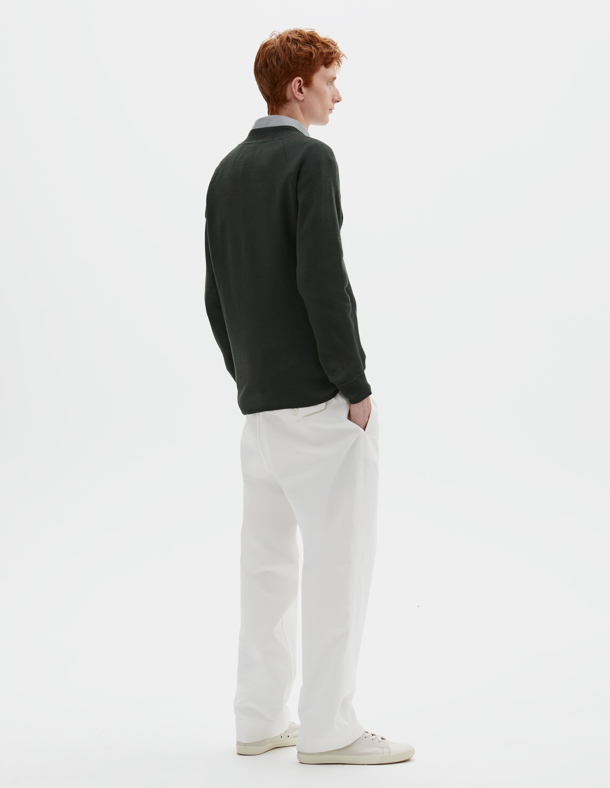 Full body image, back view of model wearing dark grey merino wool sweatshirt with raglan sleeve and contrast rib on neck, cuffs and hem. Styled over grey shirt, with white trousers and beige canvas trainers.