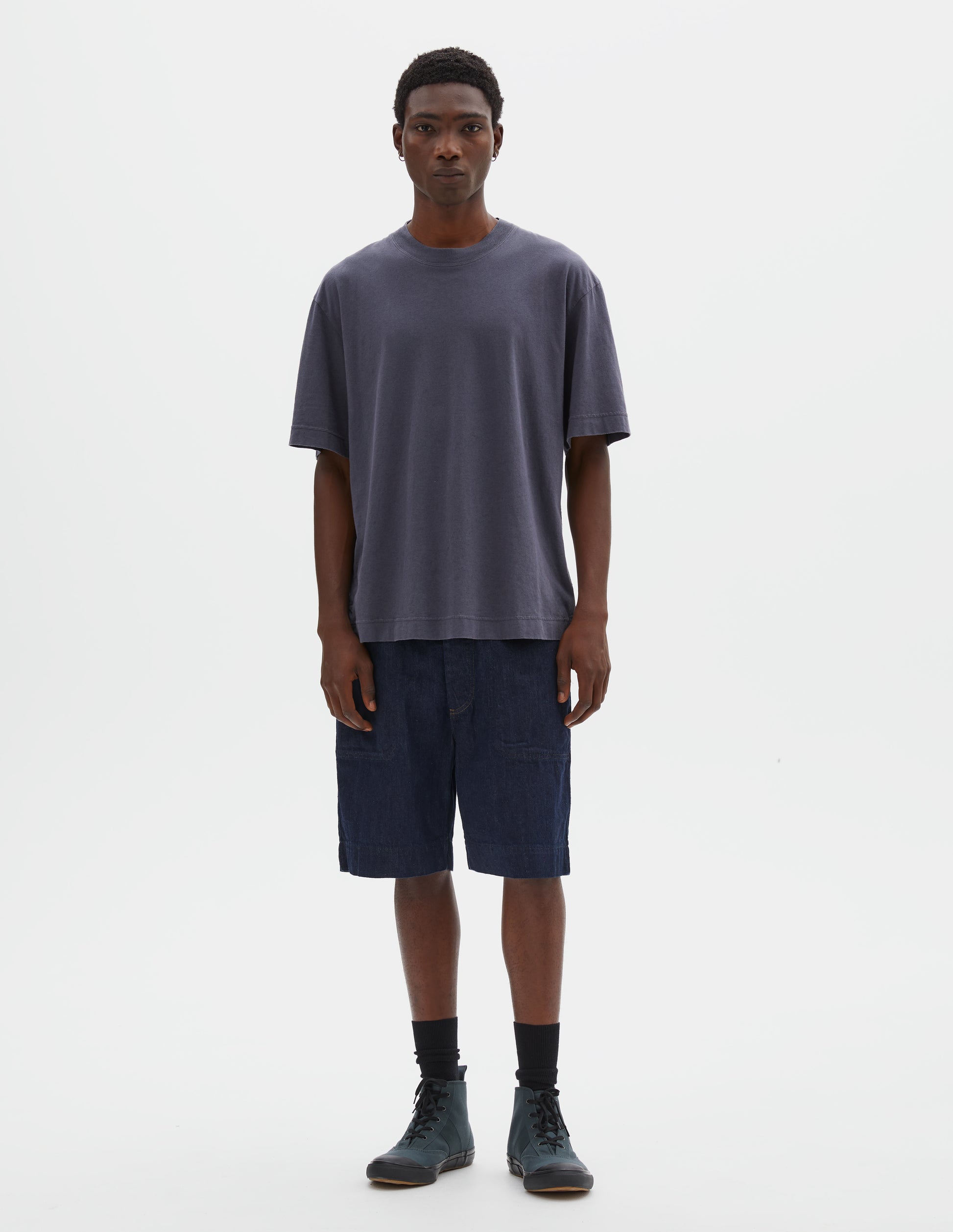 Full body image, front view of model wearing greyish blue cotton linen short sleeve t-shirt with vent split hem and herringbone tape detailing inside the neck. Styled with indigo blue denim shorts, black socks and blue canvas high top trainers.