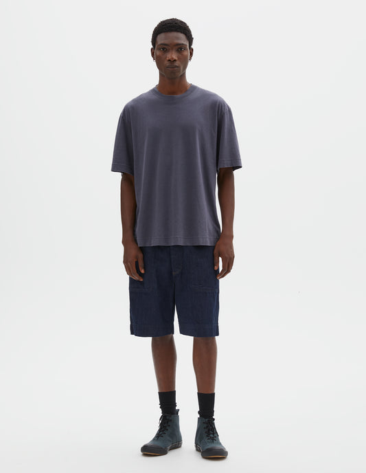 Full body image, front view of model wearing greyish blue cotton linen short sleeve t-shirt with vent split hem and herringbone tape detailing inside the neck. Styled with indigo blue denim shorts, black socks and blue canvas high top trainers.