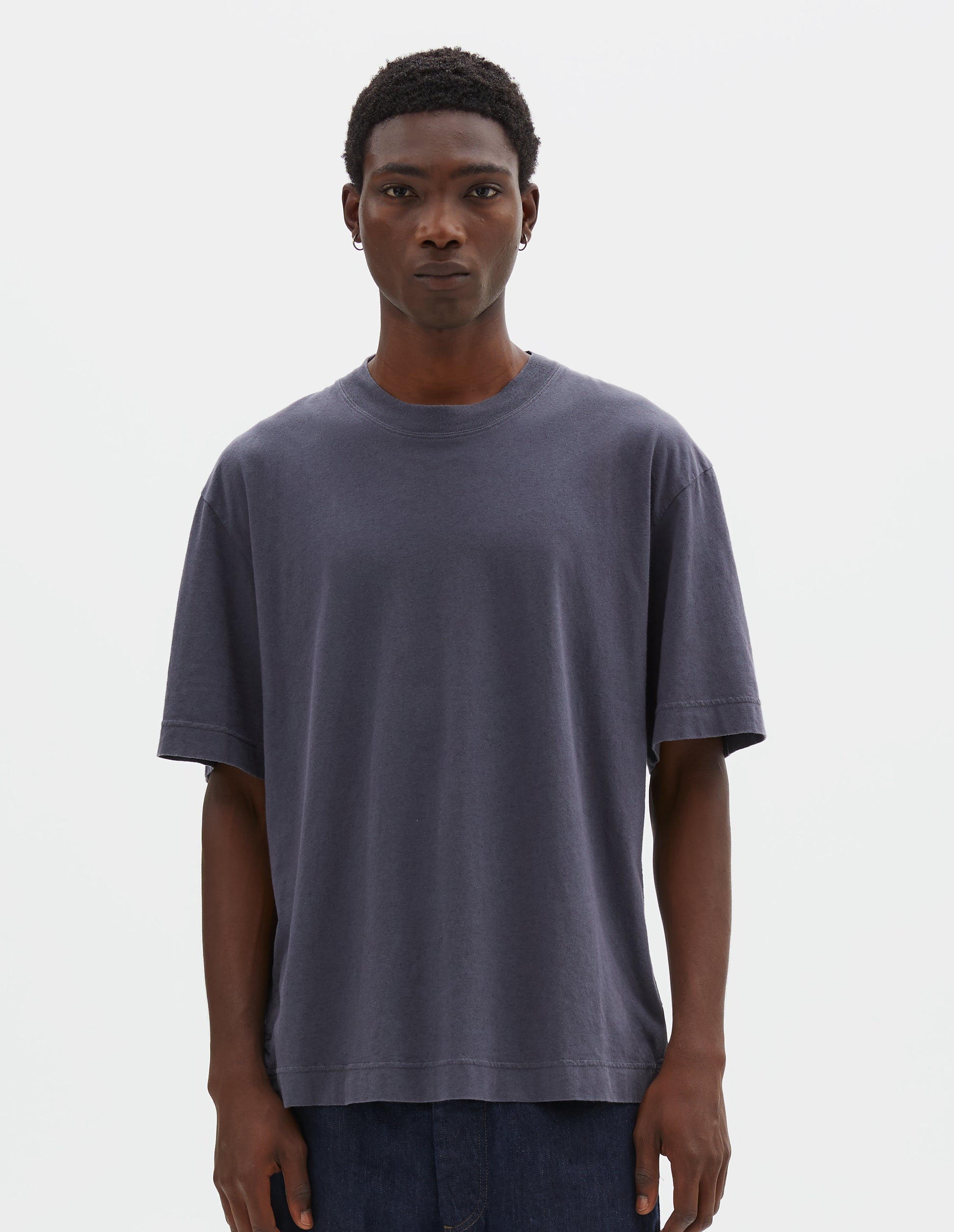 Cropped body image, front view of model wearing greyish blue cotton linen short sleeve t-shirt with vent split hem and herringbone tape detailing inside the neck. Styled with indigo blue denim shorts.