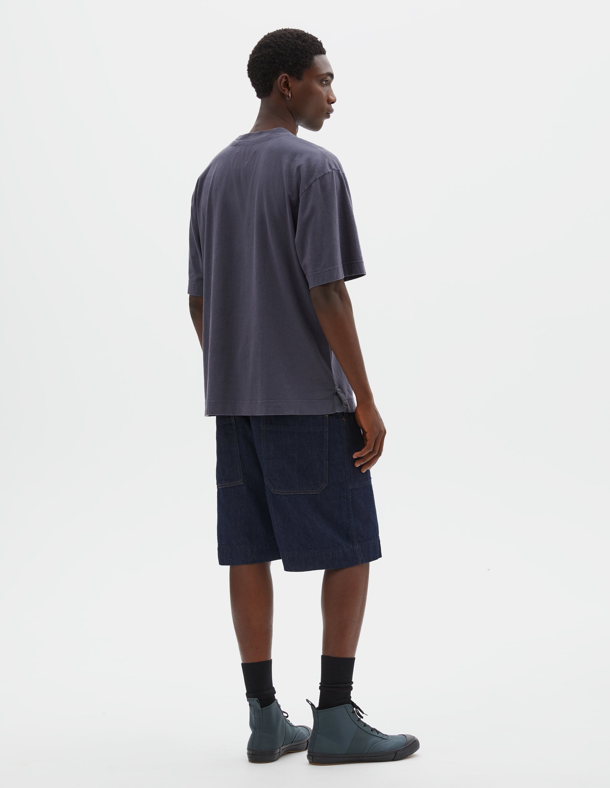 Full body image, back view of model wearing greyish blue cotton linen short sleeve t-shirt with vent split hem. Styled with indigo blue denim shorts, black socks and blue canvas high top trainers.