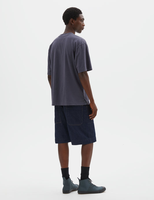 Full body image, back view of model wearing greyish blue cotton linen short sleeve t-shirt with vent split hem. Styled with indigo blue denim shorts, black socks and blue canvas high top trainers.