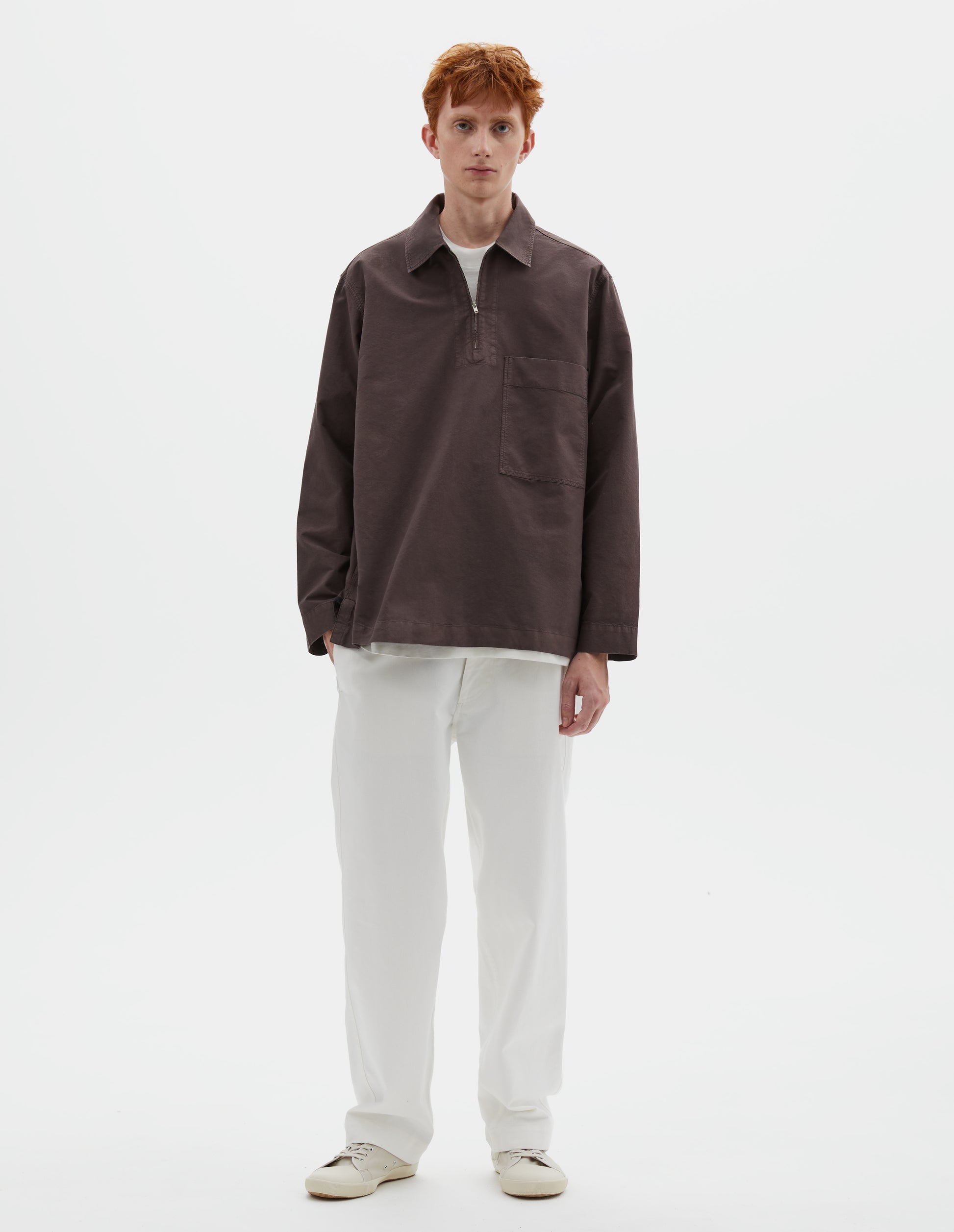 Full body image, front view of model wearing relaxed brown shirt with wide stitch-through placket with half zip, large patch chest pocket, straight cuffs and and side split hem. Styled with white trousers and beige trainers.