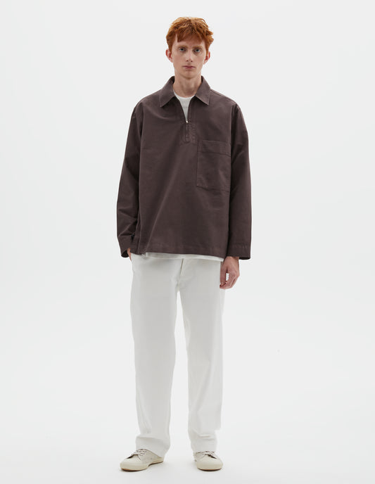 Full body image, front view of model wearing relaxed brown shirt with wide stitch-through placket with half zip, large patch chest pocket, straight cuffs and and side split hem. Styled with white trousers and beige trainers.