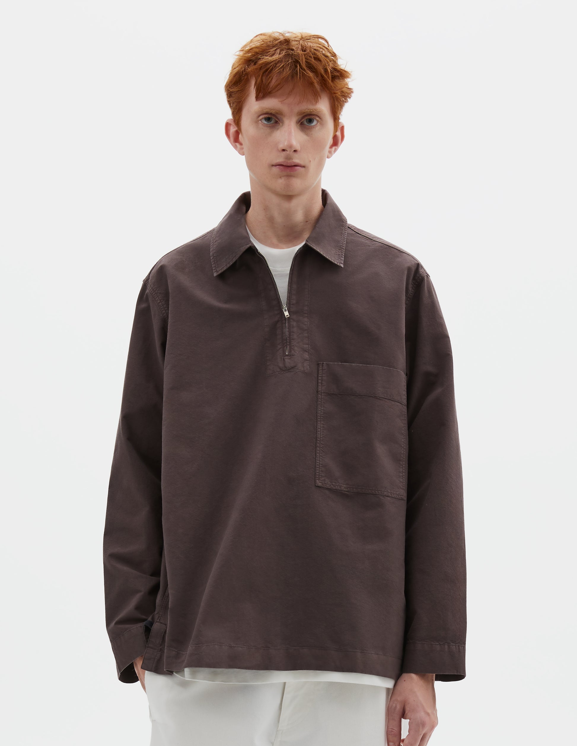 Cropped body image, front view of model wearing relaxed brown shirt with wide stitch-through placket with half zip, large patch chest pocket, straight cuffs and and side split hem. Styled over white t-shirt, and with white trousers.