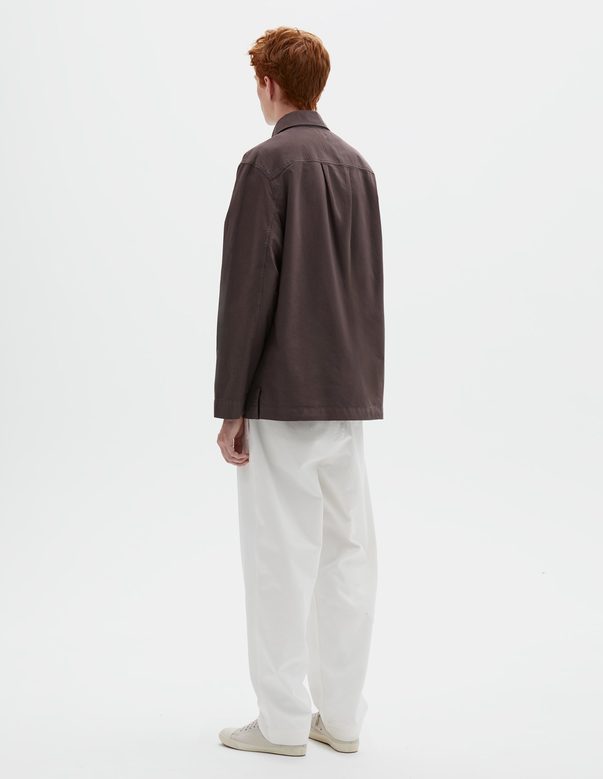 Full body image, back view of model wearing relaxed brown shirt with straight cuffs, side split hem and box pleat on back yoke. Styled with white trousers and beige trainers.
