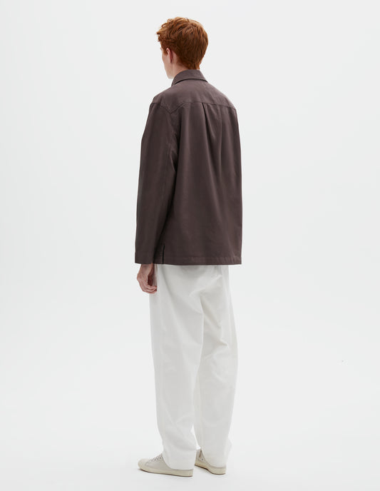 Full body image, back view of model wearing relaxed brown shirt with straight cuffs, side split hem and box pleat on back yoke. Styled with white trousers and beige trainers.