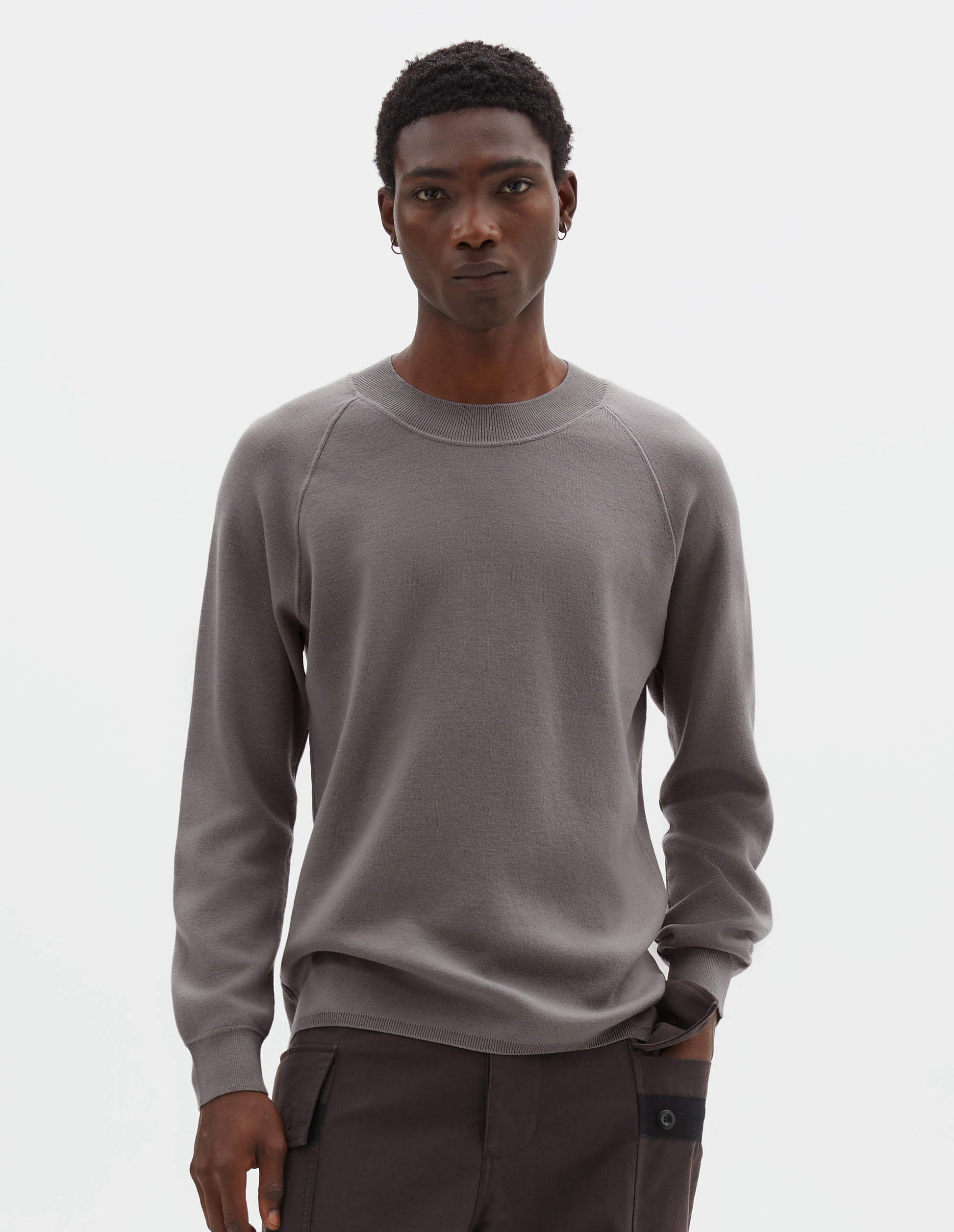 Cropped body image, front view of model wearing light grey merino wool sweatshirt with raglan sleeve and contrast rib on neck, cuffs and hem. Styled with reddish brown trousers.