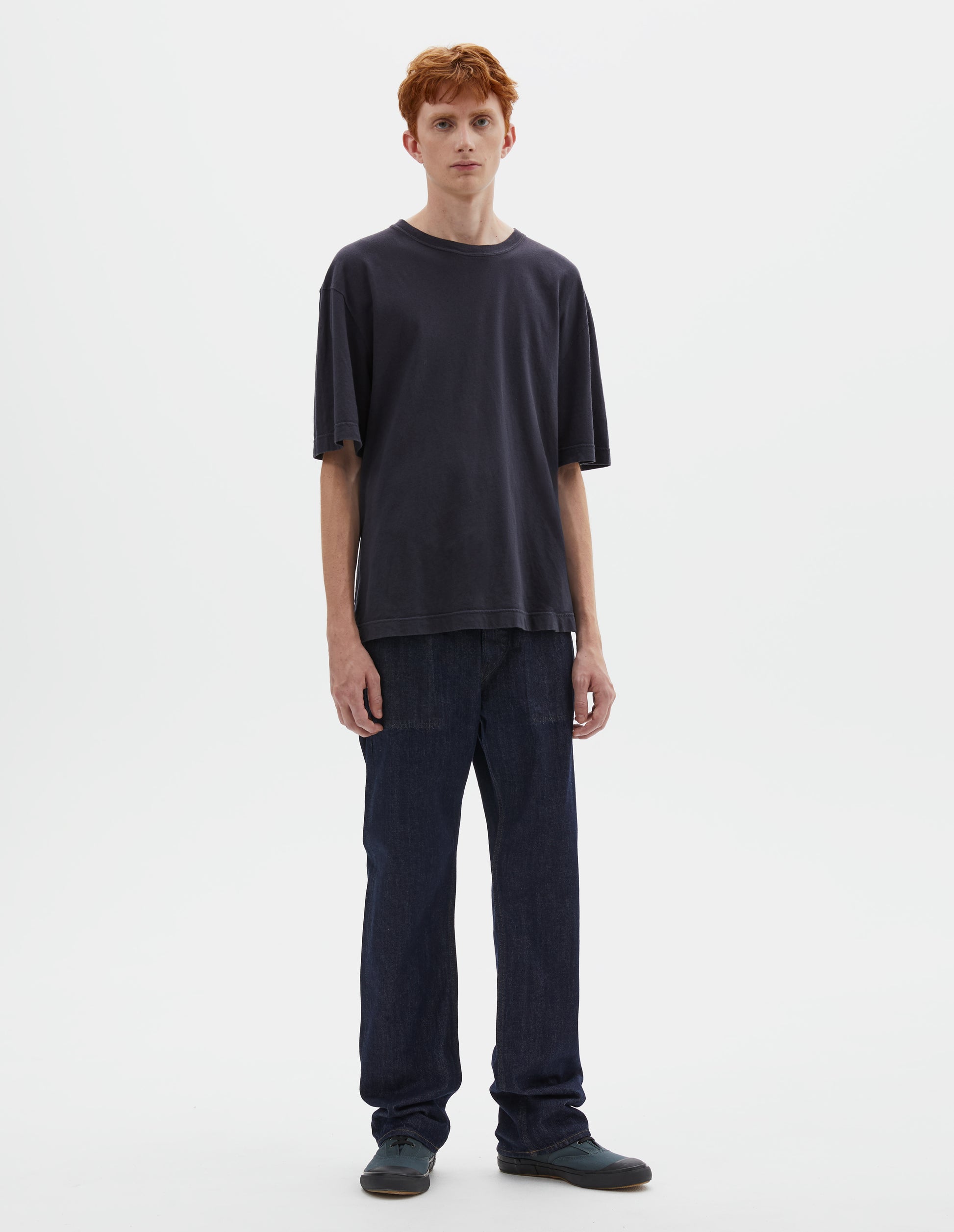 Full body image, front view of model wearing boxy dark blue t-shirt with herringbone tape detailing on inside neck and vent split hem. Styled with indigo blue jeans and blue canvas trainers.