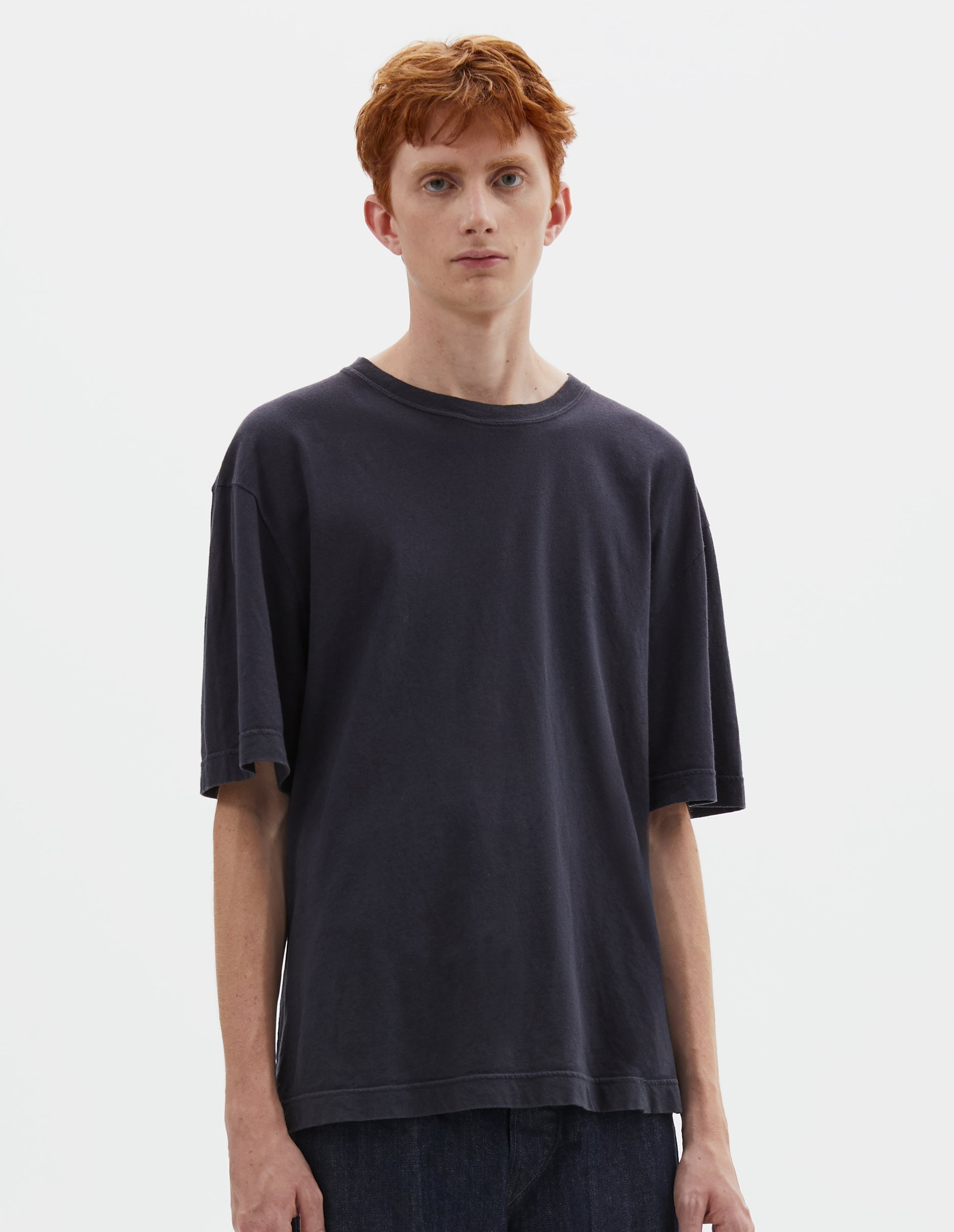 Cropped body image, front view of model wearing  boxy dark blue t-shirt with herringbone tape detailing on inside neck and vent split hem. Styled with indigo blue jeans.