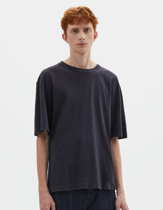 Cropped body image, front view of model wearing  boxy dark blue t-shirt with herringbone tape detailing on inside neck and vent split hem. Styled with indigo blue jeans.