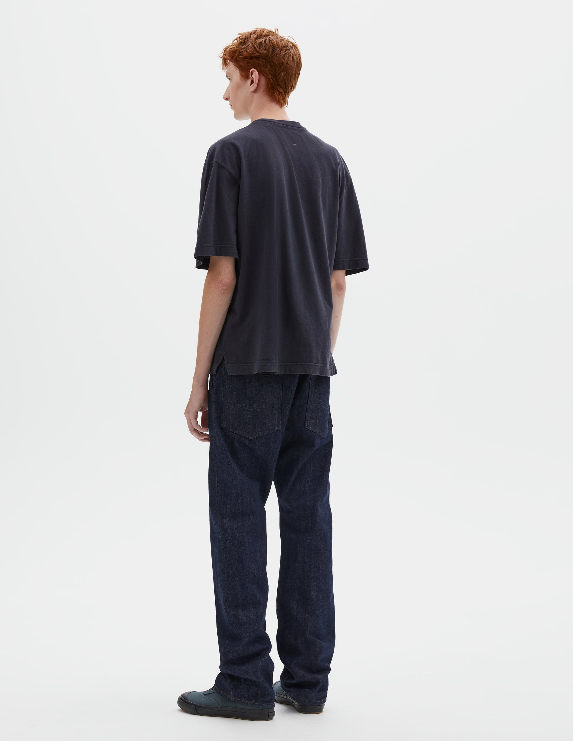 Full body image, back view of model wearing boxy dark blue t-shirt with vent split hem. Styled with indigo blue jeans and blue canvas trainers.