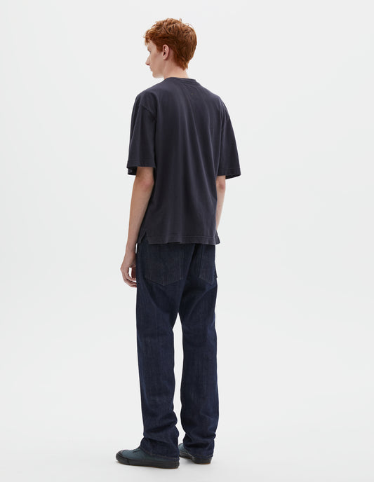 Full body image, back view of model wearing boxy dark blue t-shirt with vent split hem. Styled with indigo blue jeans and blue canvas trainers.
