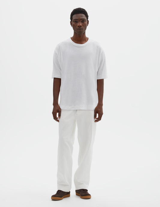 Full body image, front view of model wearing boxy white t-shirt with herringbone tape detailing on inside neck and vent split hem. Styled with off white trousers and brown suede trainers.